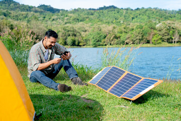 Asian man sit on grass field and use mobile phone that get power from solar cells during travel and camping in national park.