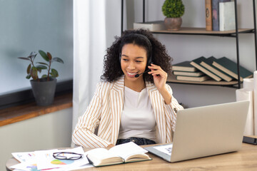 Fototapete - Successful African American woman entrepreneur or office worker in headset using a laptop for working remotely, communicates with employees online sitting at the desk in the office