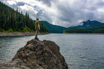 Adventurous athletic male hiker standing on a large bolder on the shore of an alpine lake on a cloudy day in the Pacific Northwest.
