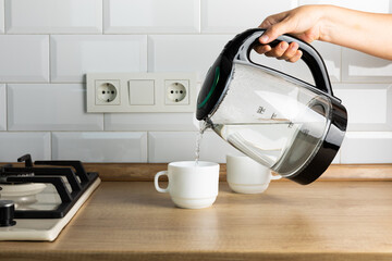 A man brews tea using boiling water from an electric kettle in the kitchen at home.Kettle for boiling water and making tea and coffee.Home appliances for hot drinks.Side view, space for text.