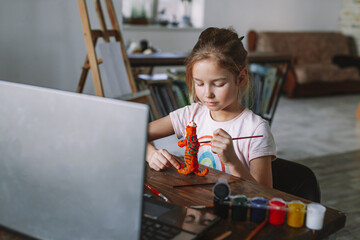 The beautiful cute girl creates crafts at home from the video lesson. A cheerful child is engaged in a hobby on a laptop. Elementary school student doing homework at homeschooling via video link.