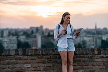 Wall Mural - a portrait of beautiful girl tourist standing leaned on a brick fence of old fortress. she is happy about visiting new place, holding paper map and cup of coffee. 