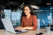 © Liubomir - Happy and smiling hispanic businesswoman typing on laptop, office worker with curly hair and glasses happy with achievement results, at work inside office building.