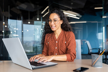 Wall Mural - Happy and smiling hispanic businesswoman typing on laptop, office worker with curly hair and glasses happy with achievement results, at work inside office building.
