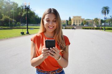 Wall Mural - Brazilian young woman laughing and texting on her mobile phone walking outdoors on sunny day