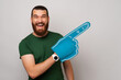 Wide smiling man is wearing blue fan glove and pointing aside with it over grey background.