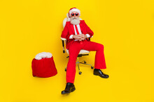 Photo Of Serious Confident Man Pensioner Dressed Red Suit Santa Hat Sitting Chair Preparing Gifts Isolated Yellow Color Background