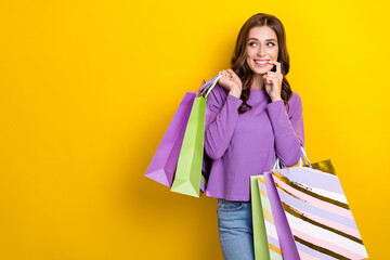 Sticker - Portrait of cheerful cunning tricky girl hold bags finger touch lip look empty space isolated on yellow color background