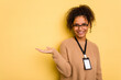 © Asier - Young brazilian woman with a badge isolated on yellow background showing a copy space on a palm and holding another hand on waist.