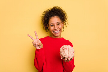 Young brazilian woman holding a brain model isolated joyful and carefree showing a peace symbol with fingers.