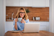 © sepy - Smiling middle aged woman using laptop and having video call while working from home. Confident female drinking tea while sitting at table at the kitchen.