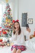 © StockPhotoRepublic - Cute beautiful young asian lady woman wearing reindeer headband selfie and video call with mobile phone posing in front a big Christmas tree with lots of decoration lights gift box and ornaments