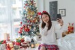 © StockPhotoRepublic - Cute beautiful young asian lady woman wearing reindeer headband selfie and video call with mobile phone posing in front a big Christmas tree with lots of decoration lights gift box and ornaments