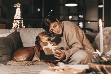 Happy Woman With Dog Cuddling And Unpacking Gifts, Looking Excitedly Into The Box, In A Cozy Environment At Home On The Couch, Mysterious, Warm Colors