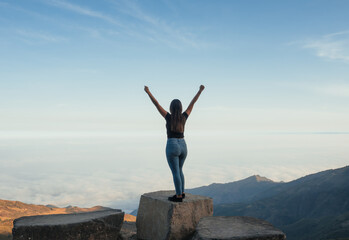 woman standing on a rock with a landscape full of sky and mountains, in the Ecuadorian highlands, she raises her hands as a sign of success for having achieved her goals.