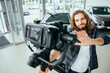 © Iryna - Social media influencer creating content. man shooting video of himself using camera on tripod. Smiling bearded hipster long haired guy communicating with subscribers in automobile showroom.