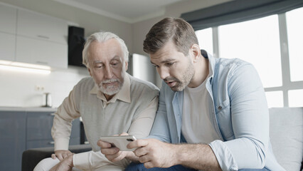 Wall Mural - Happy young man and senior father looking through photos on smartphone together, fun