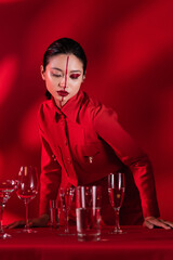 Wall Mural - asian woman with bright makeup on face divided with line posing near glasses of water on red background with shadow.