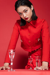 Wall Mural - brunette asian woman in elegant jacket standing near glasses with clean water on red background.