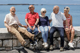 Fototapeta Miasto - gruppo di anziani amici seduti nel muretto di un porto di mare, si rilassano chiacchierando felici.