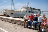 Fototapeta Uliczki - gruppo di anziani amici seduti nel muretto di un porto di mare, si rilassano chiacchierando felici.