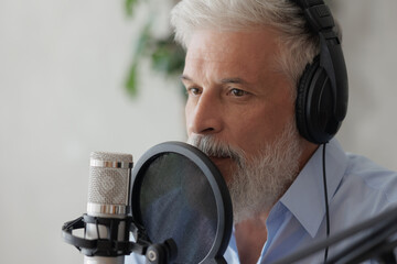 older man with gray hair irecording podcast in recording studio with microphone and headphones. senior radio presenter or interviewer. recording audio content for social media online