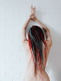 A naked girl with long dreadlocks in her hair stands near a white brick wall with her hands up