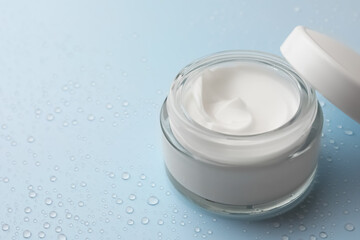 Jar of face cream on light blue surface covered with water drops, space for text