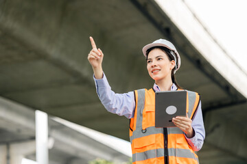 Wall Mural - Asian engineer worker woman or architect looking construction with tablet wear white safety helmet in construction site. Standing at highway concrete road site. Progress planning of highway bridge.