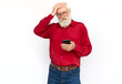 Disappointed senior man using phone with hand on head. Tired Caucasian male model with gray hair and beard in red shirt and glasses looking at screen, covering forehead. Modern technology concept