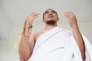 Wall Mural - Asian muslim man wearing white ihram clothes and praying while holding prayer beads