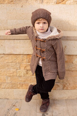 Wall Mural - a boy in knitted things a hat and a brown jacket is standing on the street leaning against the wall