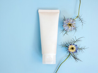 White squeeze cosmetic cream tube and blue Nigella flowers on blue background. Mockup moisturizer blank bottle for branding - skincare, cream, cleanser, make-up removal, gel, sunscreen. Front view