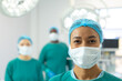 © wavebreak3 - Portrait of smiling biracial female surgeon in mask and cap in operating theatre with colleagues