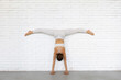 Young woman trainer practicing yoga doing Adho Mukha Vrikshasana exercise with Samakonasana, handstand with transverse splits with legs against the wall, inverted asana, exercising in white clothes