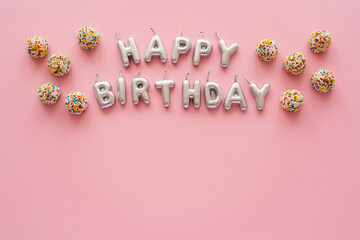 Top view of candles in shape of Happy Birthday lettering near colorful sweets on pink background.