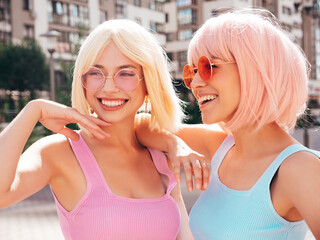 Two young beautiful smiling hipster female in trendy summer clothes. Sexy carefree women posing in the street in wigs. Positive funny models having fun at sunset. In sunglasses. At sunny day