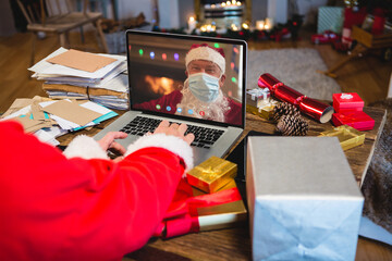 Sticker - Santa claus with christmas presents having video call with caucasian santa claus with face mask