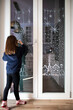 © silvia - Little caucasian child girl decorating home window for Christmas with white pencil (liquid chalk). Festive home decor idea. Vertical shot.