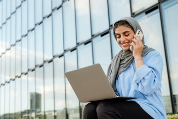 Poster - Cheerful muslim woman talking on cellphone and using laptop while sitting outdoors