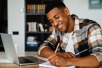 Canvas Print - Young african man working on laptop and writing down notes while sitting at table at home