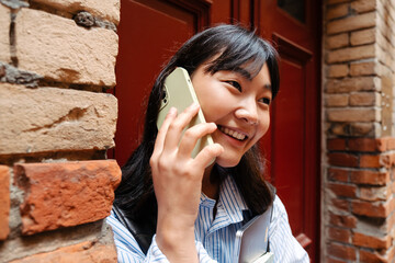 Wall Mural - Young asian girl talking on mobile phone and holding laptop outdoors