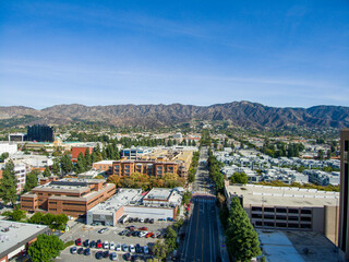 Wall Mural - aerial shot of office buildings, apartments and shops surrounded by lush green trees, cars driving and parked on the street with majestic mountains and a clear blue sky in Burbank California USA