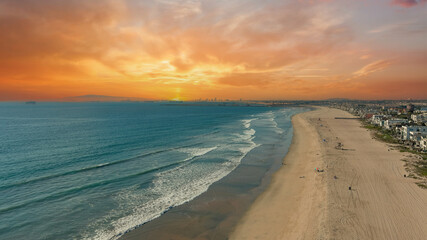 Wall Mural - aerial shot of the coastline with blue ocean water and homes along the sand on the beach, cars driving on the street and powerful clouds at sunset in Huntington Beach California USA