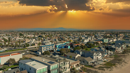 Wall Mural - an aerial shot of the vast miles of homes and lush green trees in the city skyline with cars driving on the street, majestic mountains and powerful clouds at sunset in Huntington Beach California USA