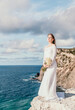 © panophotograph - Romantic bride, blond girl in white wedding dress with open shoulders posing with sea and rocks in background. Stylish young woman standing on cliff's edge and looking into a ocean mountains at sunset