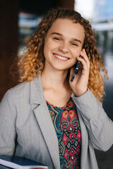 Smiling woman talking on mobile phone, standing outside building with book and laptop in her hand. Attractive beautiful girl. People lifestyle. Mobile phone