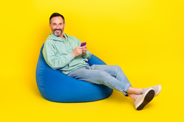 Sticker - Full length photo of positive cheerful guy dressed green hoodie sitting bean bag chatting instagram twitter telegram isolated yellow color background