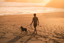 Silhouette Of A Man Walking With A Dog On The Beach At Sunset.