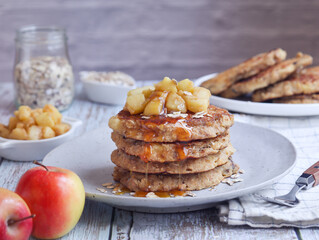 A plate of healthy rolled oat and apple pancakes topped with cooked diced apple pieces with sweet syrup. Its fruit and fork in scene with a bottle of oat grains in background 